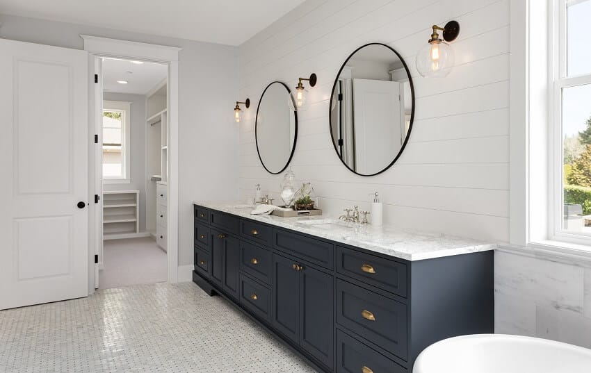 Главная ванная комната в роскошном доме с двумя раковинами, темно-синими шкафами, двумя круглыми зеркалами и видом на гардеробную