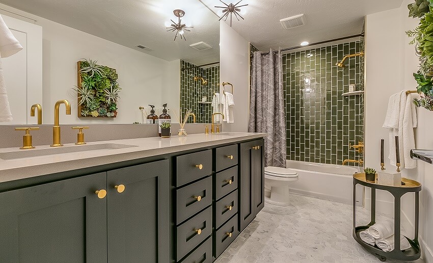 Подвальная ванная комната с зеленой плиткой в ​​душевой зоне, латунные смесители и светильники, полы из шестиугольной плитки, большое зеркало, зеленые шкафы и ящики.