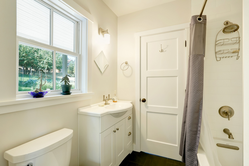 Небольшой шкаф, скрывающий трубы раковины в ванной комнате, белые стены, двери, окна, душевая зона