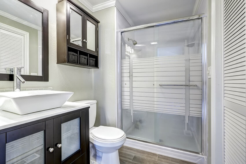 Современный интерьер ванной комнаты со стеклянной дверью, водостойкой краской для душа, коричневой тумбой с белой раковиной и зеркалом