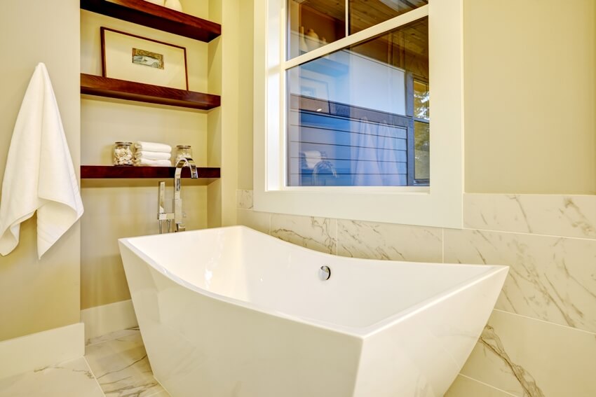 Изысканный уголок для ванной комнаты с отдельно стоящей ванной, расположенной под окном, стенной нишей с полками и белым и серым мрамором, выложенным плиткой на полу и полустене.