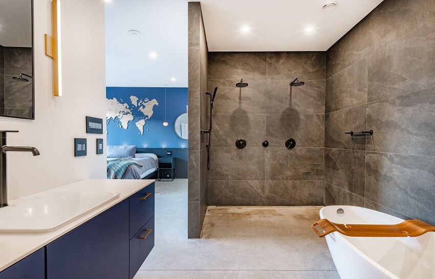 Большая ванная комната открытой планировки с отдельно стоящей ванной, двойным душем, стенами из темно-серой плитки и синими шкафчиками.